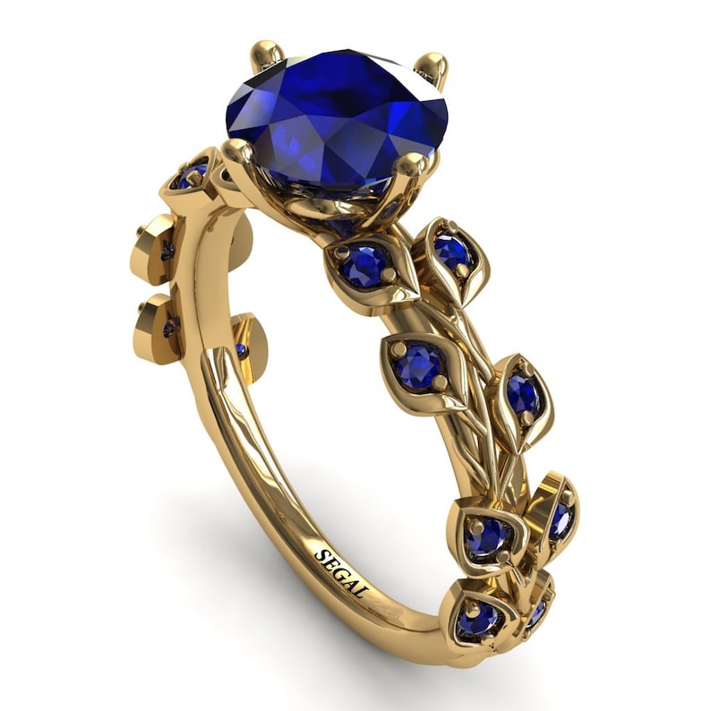 May include: A gold engagement ring with a large blue sapphire center stone and smaller blue sapphires set in leaf-shaped accents. The ring is engraved with the word "SEGAL".
