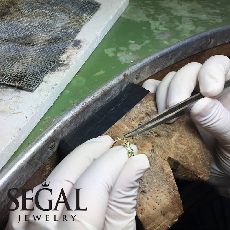May include: A close-up of a jeweler setting green gemstones into a gold band. The ring is being held with tweezers and the jeweler is wearing white gloves. The text "SEGAL JEWELRY" is visible in the lower left corner of the image.