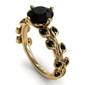 May include: A gold engagement ring with a black diamond center stone and black diamond accents on the band. The band is designed with a leaf motif. The ring is engraved with the word "SEGAL".