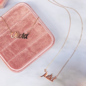 Puede incluir: Dos collares dorados con nombres. Un collar deletrea "Olivia" en una fuente cursiva, descansando sobre una caja de joyer&iacute;a de terciopelo rosa. El otro collar deletrea "Julia".