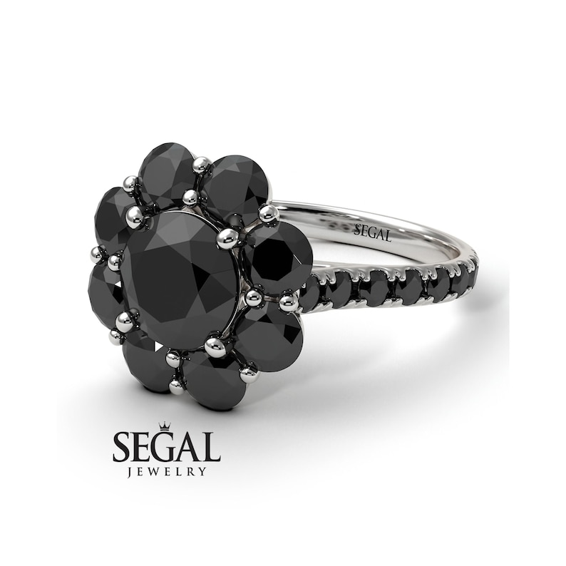 May include: A black diamond engagement ring with a flower-shaped setting. The ring is set in white gold and features a large round black diamond in the center, surrounded by smaller black diamonds. The band is also set with black diamonds.