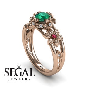 Könnte beinhalten: Ein Verlobungsring aus Roségold mit einem grünen Smaragd, der von kleineren Diamanten umgeben ist. Der Ring hat ein zartes Blumenmuster mit einem Rubin-Akzent.