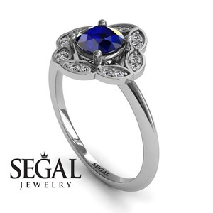 May include: A silver engagement ring with a blue sapphire center stone and a halo of smaller diamonds. The ring has a floral design and is engraved with the word "SEGAL".