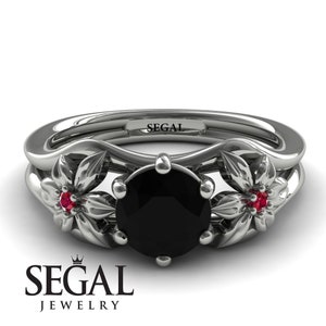 May include: A black diamond engagement ring with two ruby accents set in a floral design. The ring is made of white gold and has the inscription "SEGAL" on the band.