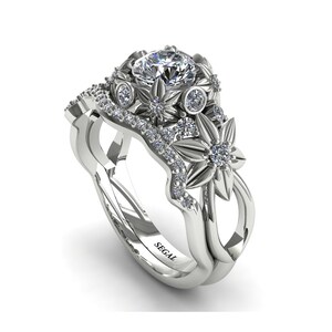 May include: A white gold engagement ring set with a large round center stone and smaller diamonds set in a floral design. The band is also decorated with smaller diamonds. The ring is engraved with the word "SEGAL".