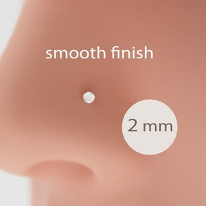 Op de afbeelding: Een kleine, zilveren neusstud met een gladde afwerking, getoond op de neus van een persoon. De stud heeft een diameter van ongeveer 2 mm. De tekst "smooth finish" staat boven de stud.
