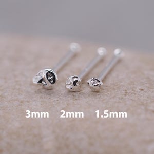 Puede incluir: Tres piercings de nariz plateados con diseños de calaveras. Los tamaños son 3 mm, 2 mm y 1,5 mm. Los piercings están sobre una superficie marrón claro.