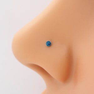 Peut inclure: Gros plan sur un piercing de nez en forme de clou avec une pierre bleue. Le clou est serti dans un cadre argenté et est porté à la narine gauche.