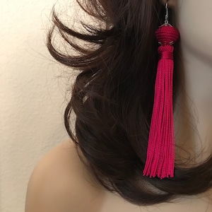 May include: A pair of long, bright pink tassel earrings with silver metal tops. The earrings are dangling from the earlobes of a person with long, dark brown hair.