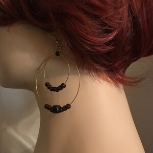 May include: Gold hoop earrings with two tiers of black wooden beads. The top tier has smaller beads and the bottom tier has larger beads, including one larger, square-shaped bead.