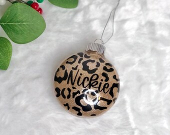 May include: A round Christmas ornament with a gold glitter base and black leopard print spots. The name "Nickie" is written in black script across the front. A silver string is attached for hanging.