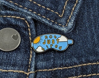 May include: A blue enamel pin with the words "not just tired" in a whimsical font. The pin features a sun, clouds, and a sleep mask design. The pin is attached to a denim jacket.