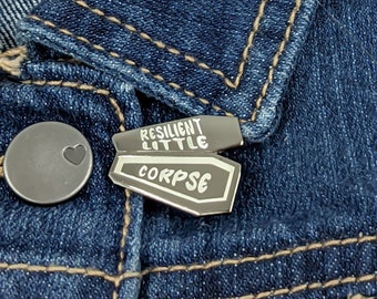 May include: A silver enamel pin with a black coffin shape. The coffin has the words "Resilient Little Corpse" written on it.