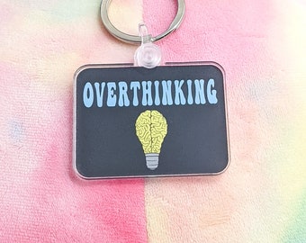 May include: A black and white keychain with the text "OVERTHINKING" in blue letters. A yellow lightbulb with a brain inside is below the text.