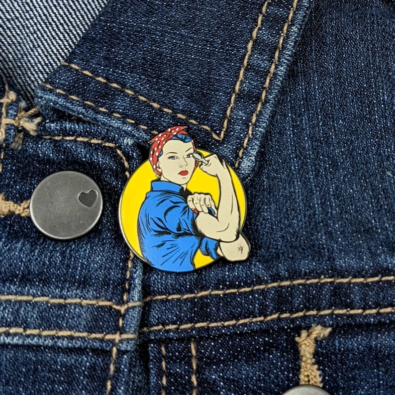 K&ouml;nnte beinhalten: Runder Emaille-Pin mit dem ikonischen Rosie the Riveter-Motiv, einer Frau, die ihren Bizeps anspannt. Der Pin hat einen gelben Hintergrund und ist auf einer dunkelblauen Jeansjacke angebracht.