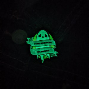Puede incluir: Un pin de esmalte verde que brilla en la oscuridad con un extraterrestre de dibujos animados y el texto "Algunas absurdeces son inevitables."