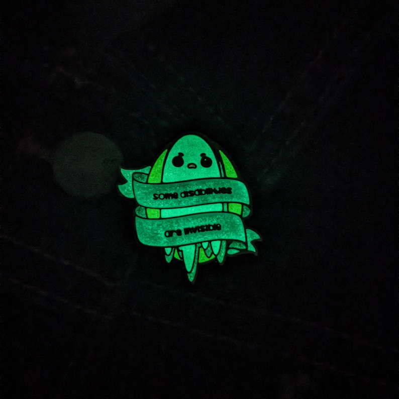 Puede incluir: Un pin de esmalte verde que brilla en la oscuridad con un extraterrestre de dibujos animados y el texto "Algunas absurdeces son inevitables."