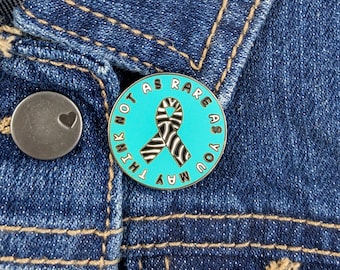 May include: A round teal pin with the text "NOT AS RARE AS YOU MAY THINK" encircling a black and white zebra-striped ribbon. The pin is attached to a denim jacket. A silver button with a heart is also visible.