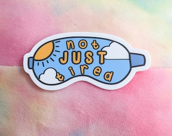 Può includere: Una maschera per dormire blu con un sole giallo e nuvole bianche. Il testo "not just tired" è scritto in arancione.