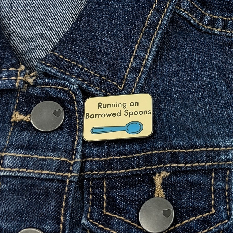 May include: A yellow enamel pin with a blue spoon and the text "Running on Borrowed Spoons" on a denim jacket.