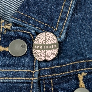 May include: A black and pink enamel pin featuring a brain with the text "the illest" on a denim jacket.