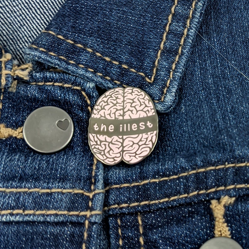 May include: A black and pink enamel pin featuring a brain with the text "the illest" on a denim jacket.