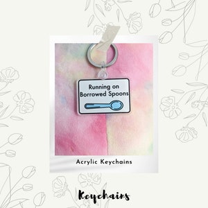 May include: A clear acrylic keychain with a white background and black outline. The keychain features a blue spoon and the text "Running on Borrowed Spoons". The keychain is attached to a silver key ring. The keychain is on a pink and yellow tie-dye background.