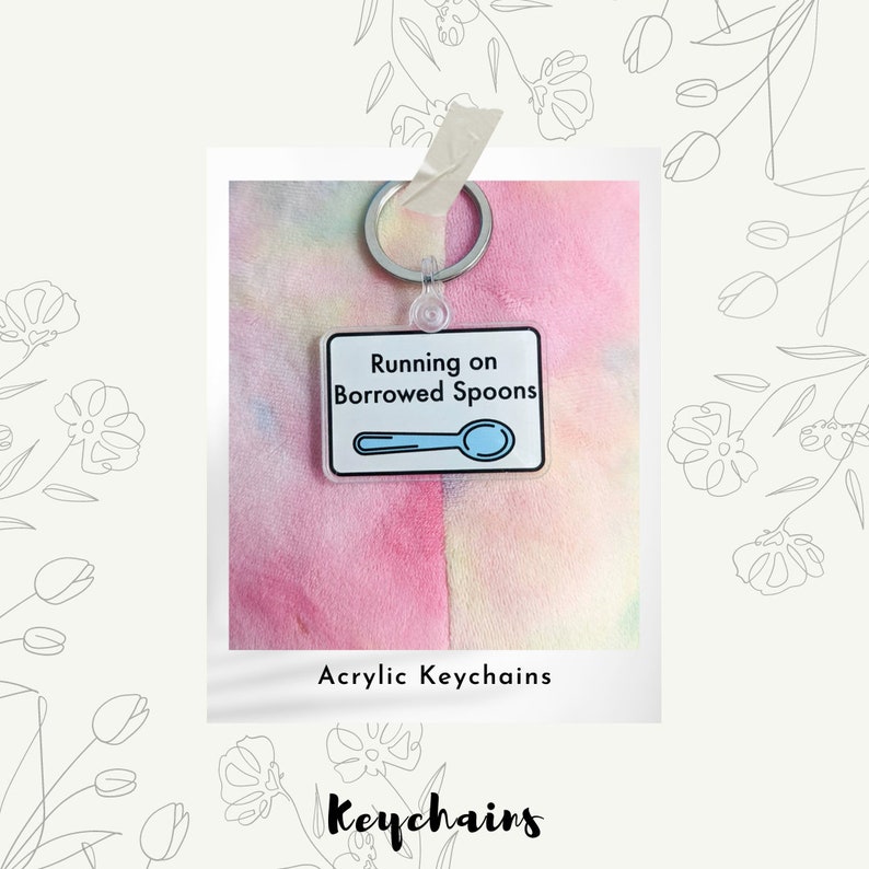 May include: A clear acrylic keychain with a white background and black outline. The keychain features a blue spoon and the text "Running on Borrowed Spoons". The keychain is attached to a silver key ring. The keychain is on a pink and yellow tie-dye background.