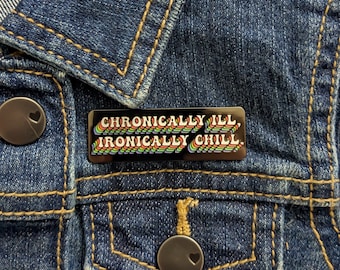 May include: A black enamel pin with the text "CHRONICALLY ILL, IRONICALLY CHILL" in rainbow-colored, wavy lettering. The pin is rectangular and set against a blue denim jacket.