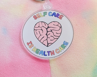 May include: A round, clear acrylic keychain with a pink brain-shaped heart in the center. The text "SELF CARE IS HEALTH CARE" is written around the heart in rainbow colors.