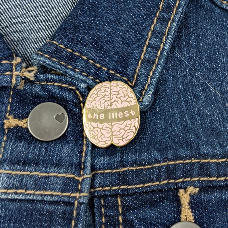 May include: Gold-colored pin with a pink brain design and the text "the illest" on a denim jacket.