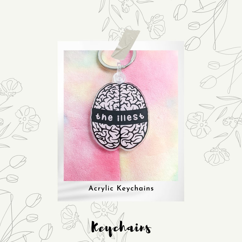 May include: A pink and white tie-dye background with a black and white brain-shaped acrylic keychain. The keychain has a black band across the middle with the text "the illest" written in white.