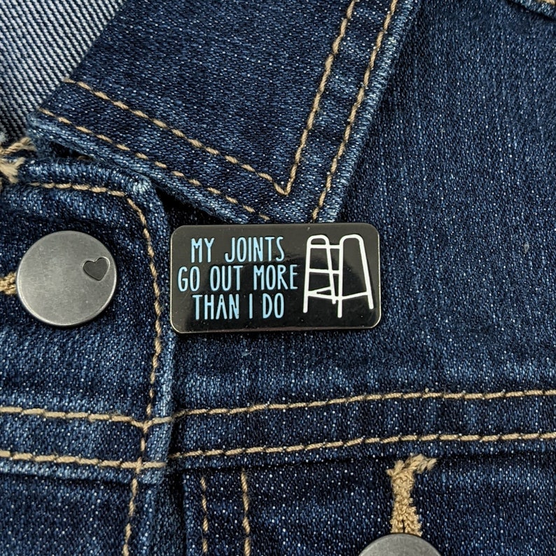 May include: A black enamel pin with a white outline of a walker and the text "My joints go out more than I do".
