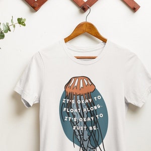 Puede incluir: Camiseta blanca con un gráfico circular azul que presenta una medusa y el texto "It's okay to float along. It's okay to just be."