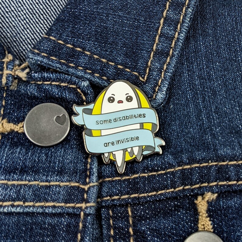 Puede incluir: Un pin de esmalte azul y amarillo con un dise&ntilde;o de fantasma. El fantasma tiene una expresi&oacute;n de sorpresa y est&aacute; rodeado de una cinta azul con el texto "Algunas discapacidades son invisibles".