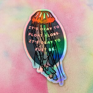 May include: A holographic sticker with a jellyfish illustration and the text "It's okay to float along. It's okay to just be."