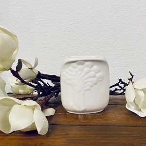 May include: A small white ceramic vase with a raised design of a tree on the front. The vase is sitting on a wooden surface with white flowers.