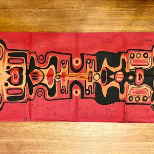 Op de afbeelding: Een rood stoffen paneel met een zwart en oranje geometrisch ontwerp. Het ontwerp heeft een herhalend patroon van gestileerde gezichten met cirkels en lijnen.