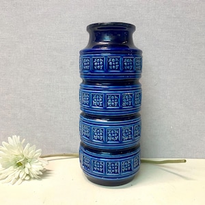 Könnte beinhalten: Eine kobaltblaue Keramikvase mit gestapeltem Design. Die Vase hat eine Reihe horizontaler Bänder mit einem sich wiederholenden Blumenmuster. Eine weiße Blume mit langem Stiel befindet sich im Vordergrund.