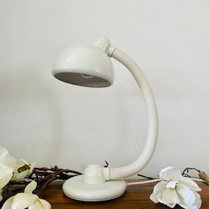 Könnte beinhalten: Eine weiße Schreibtischlampe mit einem flexiblen Schwanenhalsarm und einem weißen Schirm. Die Lampe steht auf einer Holzoberfläche mit weißen Blumen.