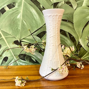 Könnte beinhalten: Weiße Keramikvase mit einem strukturierten Design. Die Vase ist hoch und schlank mit einer schmalen Öffnung. Sie steht auf einer Holzoberfläche mit einem Zweig weißer Blumen.