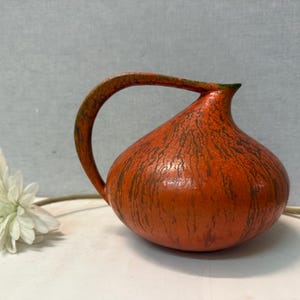 Puede incluir: Una jarra naranja decorativa con un asa curva y una superficie texturizada. La jarra tiene vetas marrones oscuras y un pequeño pico. Una flor blanca con un tallo verde está en primer plano.