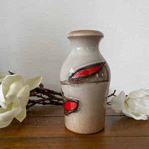 May include: A small, ceramic vase with a light brown glaze and red and black abstract designs. The vase is sitting on a wooden surface.
