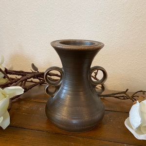 Könnte beinhalten: Eine kleine, dunkelbraune Keramikvase mit zwei Schleifenhenkeln. Die Vase hat eine leicht ausgestellte Öffnung und einen runden Körper.