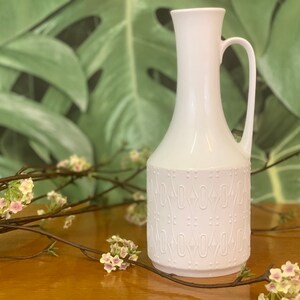 Könnte beinhalten: Weiße Keramikvase mit Henkel und geometrischem Muster. Die Vase steht auf einer Holzoberfläche mit einem Zweig rosa Blüten.