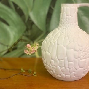 Könnte beinhalten: Weiße Keramikvase mit einer strukturierten, kieselsteinartigen Oberfläche. Die Vase hat einen runden Körper und einen Henkel.