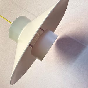 Könnte beinhalten: Eine Pendelleuchte im Retro-Stil. Die Leuchte hat einen weißen, konischen Schirm mit einem hellgrünen zylindrischen Sockel. Ein gelbes Elektrokabel hängt von oben. Die Leuchte ist an der Decke aufgehängt.