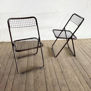 Könnte beinhalten: Zwei Vintage-Klappstühle aus Metall. Die Stühle haben ein dunkelbraunes Gestell und eine Sitzfläche und Rückenlehne mit Gittermuster. Die Stühle stehen auf einem Holzboden. Ein Stuhl ist vollständig geöffnet, der andere ist teilweise zusammengeklappt.