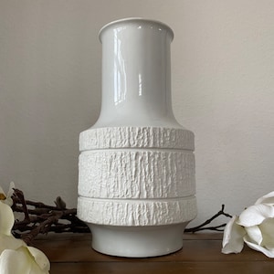 Könnte beinhalten: Eine weiße Keramikvase mit einem strukturierten, rindenartigen Design in der Mitte. Die Vase hat einen zylindrischen oberen Teil und einen abgerundeten Boden. Der Hintergrund umfasst weiße Blumen und eine Holzoberfläche.