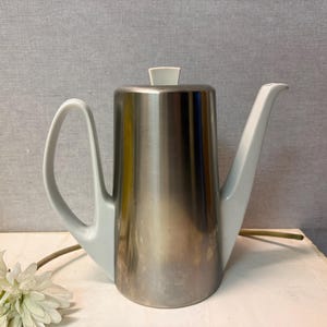 May include: A stainless steel coffee pot with a light gray handle, spout, and lid. The pot has a cylindrical body and a curved handle. The spout is long and slender. The lid is a small, rectangular shape.